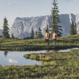 Two hikers walking across an alpine meadow at Hochkönig near Maria Alm, passing a small mountain lake with views of the impressive alpine landscape.