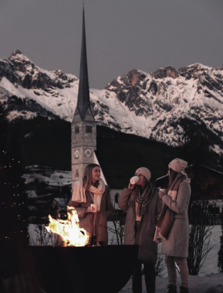 Drei Damen stehen an einer Feuerschale auf der Terrasse des Hotels die HOCHKÖNIGIN, halten Glühwein in den Händen und genießen den Ausblick auf das Steinerne Meer und die Kirche von Maria Alm