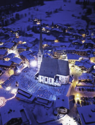 Drohnenfoto von Maria Alm im Winter mit verschneiten Dächern, der beleuchteten Kirche und stimmungsvollen Lichtern inmitten der Berglandschaft nahe dem Hotel die HOCHKÖNIGIN