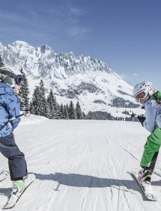 Two kids skiing joyfully on a sunny slope with a view of the Hochkönig mountains in Maria Alm.