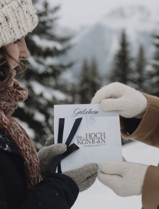 Two people exchange a HOCHKÖNIGIN gift voucher surrounded by snow and alpine scenery in Maria Alm, Austria – the perfect winter gift idea.