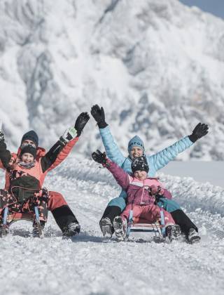 A family enjoying a fun sledding adventure in the snowy mountains of Maria Alm. Bright sunshine, fresh powder and joyful moments create the perfect winter experience for guests of the HOCHKÖNIGIN.