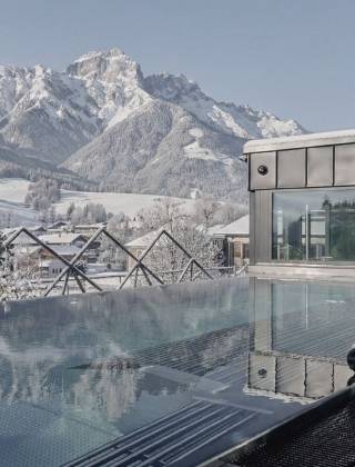 Adults-only infinity pool at the hotel die HOCHKÖNIGIN in Maria Alm with winter view of the parish church, snow-covered alpine village and the Hochkönig mountain range – luxury wellness hotel in the Salzburg region, winter holiday in Austria