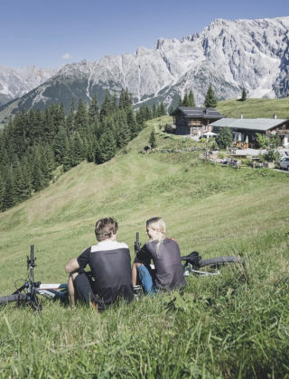 Zwei E-Biker genießen die Aussicht vor einer Berghütte am Hochkönig in Maria Alm – E-Bike Tour mit Panoramablick auf das Steinerne Meer.