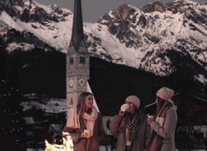 Drei Damen stehen an einer Feuerschale auf der Terrasse des Hotels die HOCHKÖNIGIN, halten Glühwein in den Händen und genießen den Ausblick auf das Steinerne Meer und die Kirche von Maria Alm