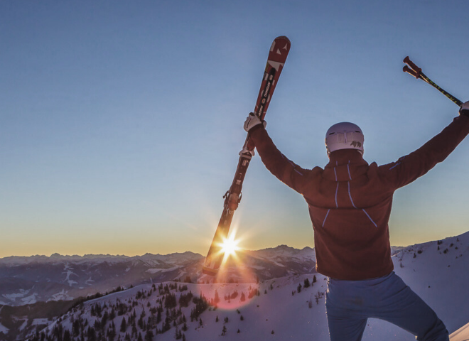 A skier raises their arms in joy at sunrise over the snowy mountains of the Hochkönig.