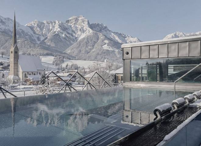 Adults-only infinity pool at the Hotel die HOCHKÖNIGIN in Maria Alm with winter views of the parish church, the snow-covered mountain village, and the Hochkönig massif – wellness hotel in the Salzburg region, winter vacation in Austria