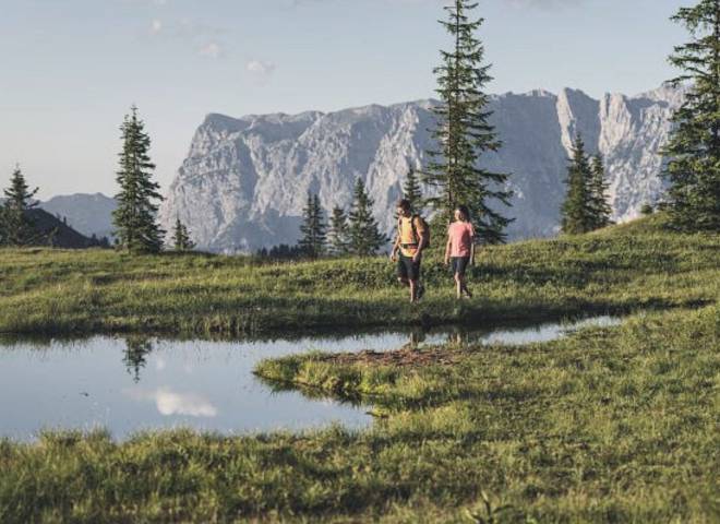 Two hikers walking across an alpine meadow at Hochkönig near Maria Alm, passing a small mountain lake with views of the impressive alpine landscape.