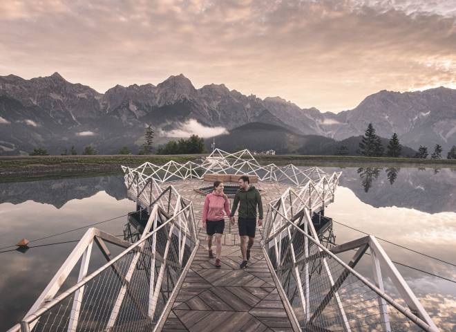 Couple walking on a wooden pier at a mountain lake at Hochkönig near Maria Alm at sunset, with mountain reflections in the water.