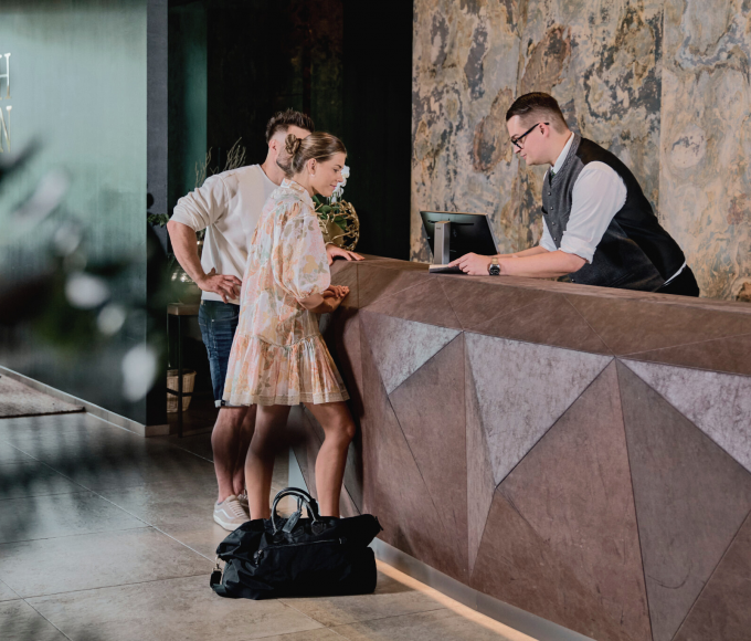 Ein junges Pärchen steht vor der stylischen Rezeption wärend ihenen ein männlicher Rezeptionist alles zu ihrem Check in erklärt