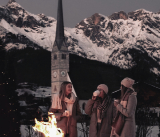 Drei Damen stehen an einer Feuerschale auf der Terrasse des Hotels die HOCHKÖNIGIN, halten Glühwein in den Händen und genießen den Ausblick auf das Steinerne Meer und die Kirche von Maria Alm