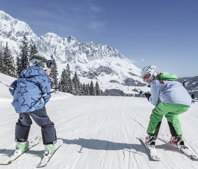 Zwei Kinder fahren lachend Ski auf der sonnigen Piste mit Blick auf das Hochkönigmassiv in Maria Alm.