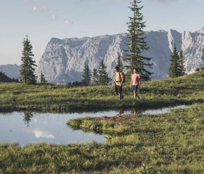 Two hikers walking across an alpine meadow at Hochkönig near Maria Alm, passing a small mountain lake with views of the impressive alpine landscape.