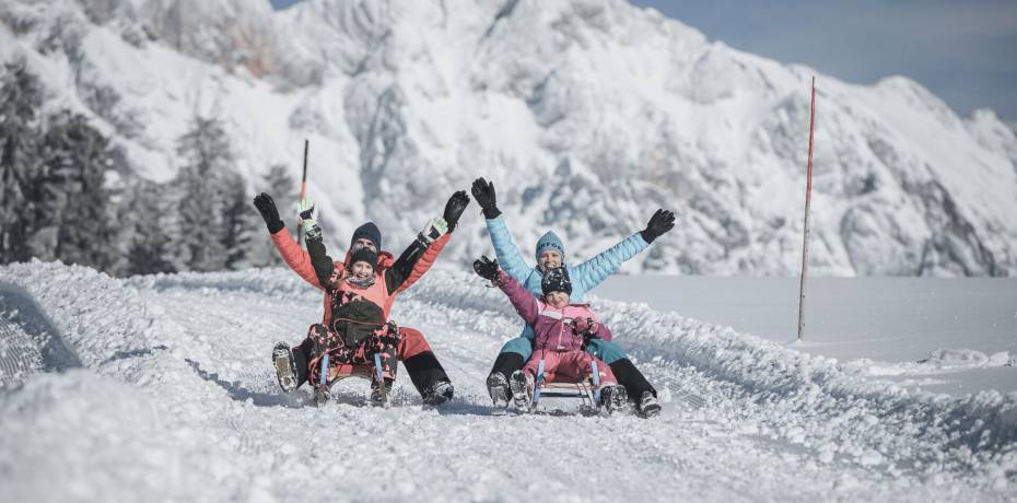 Eine Familie genießt beim Rodeln den Winterurlaub in Maria Alm. Strahlender Sonnenschein, verschneite Berglandschaft und lachende Kinder machen den Tag im Schnee zu einem unvergesslichen Erlebnis. Perfekt für Gäste, die im Hotel die HOCHKÖNIGIN aktive Wintermomente und gemeinsame Familienzeit suchen.