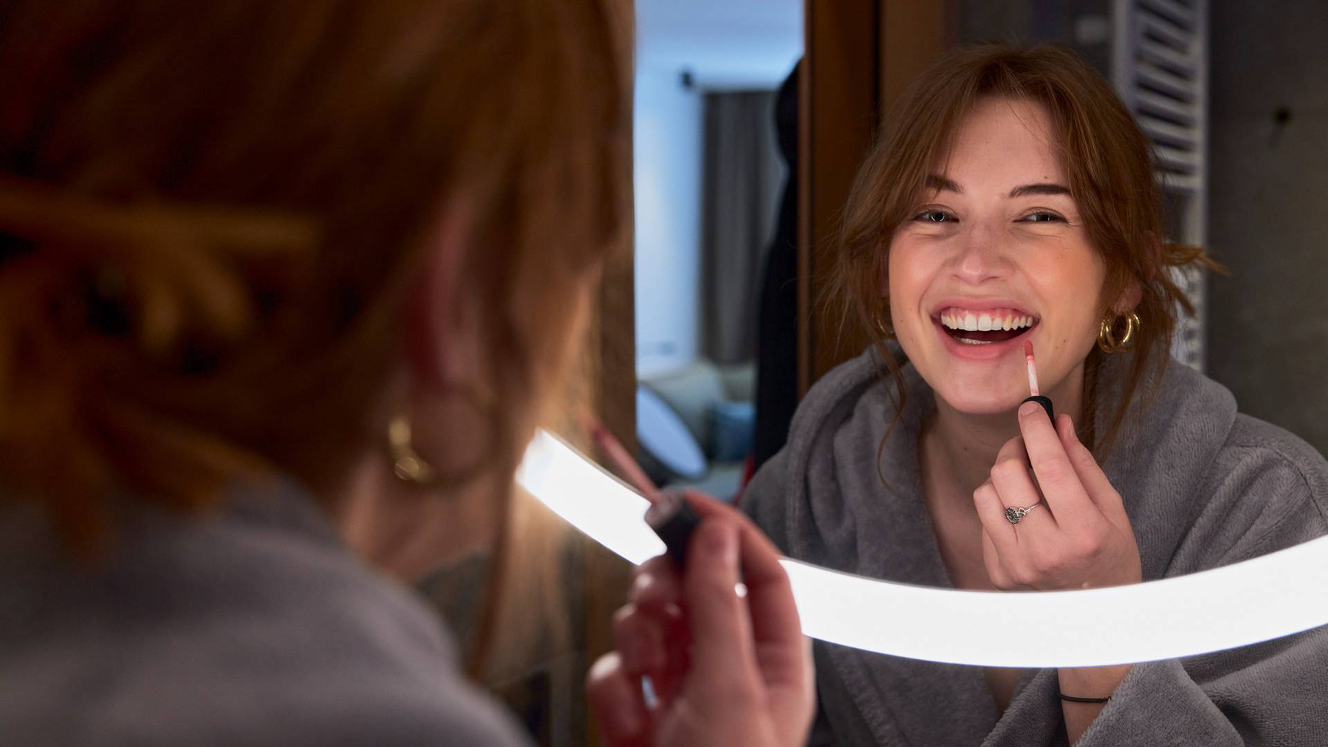 Frau trägt Lipgloss vor beleuchteten Spiegel auf und lacht dabei