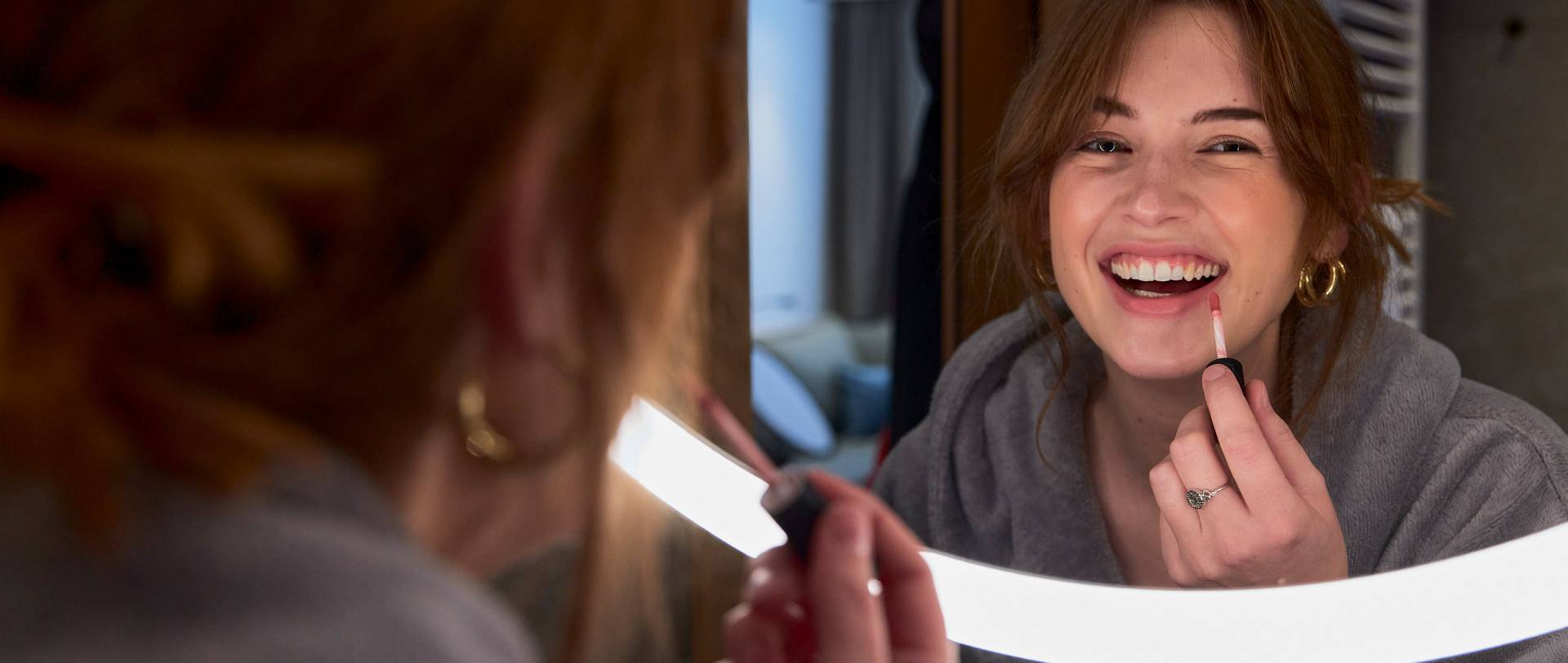Frau trägt Lipgloss vor beleuchteten Spiegel auf und lacht dabei